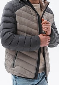 Muže, který zapíná zip na šedé a černé prošívané péřové bundě přes béžový svetr a modré džíny, s důrazem na detaily bundy.