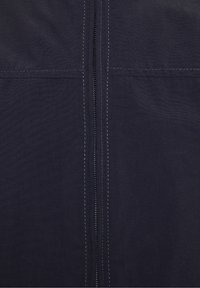 Chaqueta de combinación oscura con cremalleras, que presenta costuras externas, un cierre de cremallera vertical y una textura de tela suave y mate.