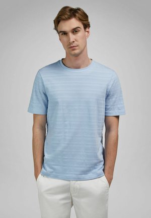 Jonge man die een lichtblauw gestructureerd shirt met korte mouwen en witte broek draagt, staat met zijn handen in zijn zakken tegen een grijze achtergrond.