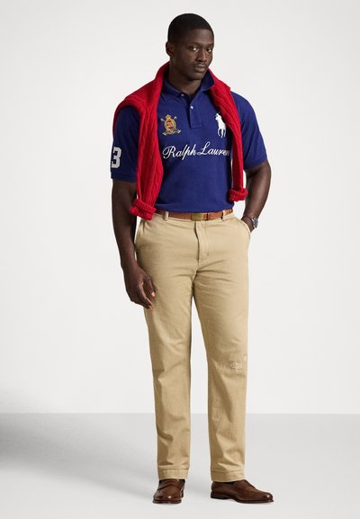 Mann iført marineblå polotrøye med broderi, rød genser drapert over skuldrene, beige bukser, brunt belte, klokke og brune skinnsko.