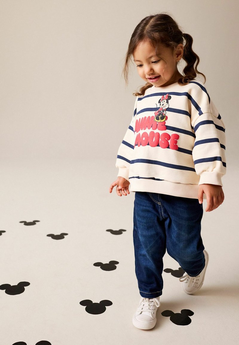 Sudadera blanca con rayas y gráfico de "Minnie Mouse" en rojo, combinada con vaqueros azules y zapatillas blancas. Siluetas negras de Mickey Mouse en el suelo.