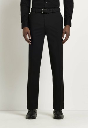 Homme portant un pantalon habillé noir, une ceinture tissée noire, une chemise habillée noire et des chaussures en cuir noir, debout devant un fond clair uni.