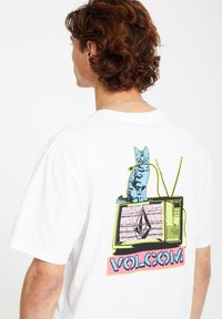 T-shirt en coton blanc avec un imprimé coloré d'un chat sur une télévision vintage, avec le texte "VOLCOM" en rose et bleu en dessous.