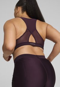 Sujetador deportivo púrpura con espalda de malla y diseño cruzado. Textura de tela suave, combinado con leggins púrpuras a juego. Detalle de logo mínimo.
