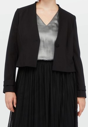 Donna che indossa un blazer nero corto sopra una camicetta in raso argento con scollo a V e una gonna nera plissettata, in piedi davanti a uno sfondo semplice.