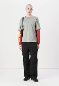 T-shirt grigio chiaro sovrapposto a una maglietta a maniche lunghe rossa, abbinato a pantaloni cargo neri larghi e scarpe nere slip-on.