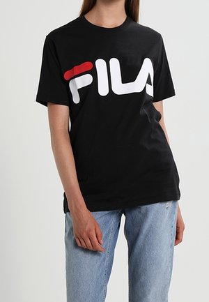 Personne portant un t-shirt noir avec un grand logo FILA blanc et rouge et un jean bleu clair sur un fond uni.