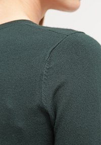 Mörkgrön stickad tröja med en närbild av axelsömmen. Tyget har en jämn textur och diskret sömnadsdetalj.