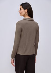 Femme aux longs cheveux noirs vue de dos, portant un haut à manches longues taupe et un pantalon noir, sur un fond clair uni.