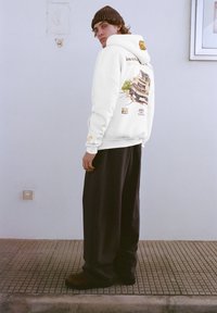 Fehér kapucnis pulóver grafikával a hátoldalán, szöveggel és színekkel. Fekete, bő szárú nadrággal és barna lábbelivel párosítva.