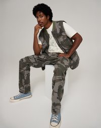 Kamuflaaživest ja -püksid roheline ja halli tooni, koos valge graafilise T-särgi ja siniste spordijalatsitega, üheltoonilisel taustal.