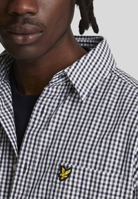 Camicia a scacchi bianca e nera con colletto, dotata di tasca sul lato sinistro del petto e un logo ricamato giallo. Materiale in cotone.