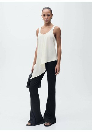 Cream asymmetric tank top paired with black flared pants, featuring a light texture. Model holds a gray woven bag, barefoot with minimal accessories.