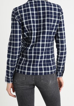 Frau trägt ein eng anliegendes marineblau-weiß kariertes Langarmhemd und dunkelgraue Jeans, von hinten gezeigt.