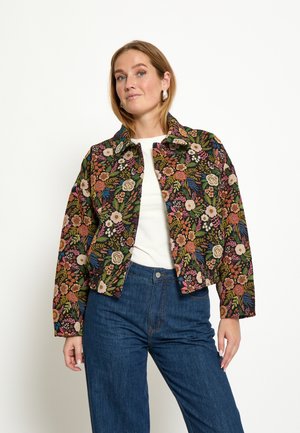 Kvinde iført en farverig blomstret jakke over en hvid top kombineret med højttaljede blå jeans, stående mod en hvid baggrund.