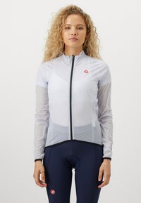 Hvit lettvekt syklingjakke med glidelås foran, nettingdesign og svarte detaljer. Båret over mørke sykkelshorts, har en merkevarelogo.