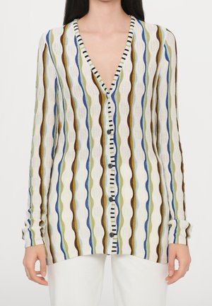 Cardigan à manches longues boutonné avec des rayures verticales ondulées en bleu, marron, vert et blanc, porté sur un pantalon blanc.