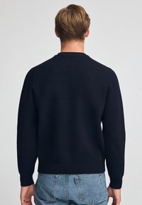 Mann trägt einen schwarzen Strickpullover und blaue Jeans, von hinten vor einem schlichten hellen Hintergrund gesehen.
