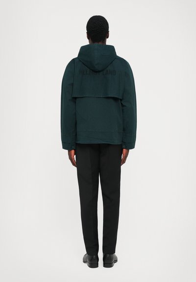 Άνδρας που φοράει σκούρο πράσινο φούτερ με κουκούλα και πλάτη και μαύρο παντελόνι, στέκεται μπροστά σε άσπρο φόντο.