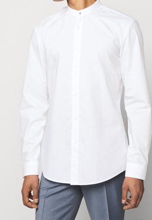 Man wearing a long-sleeve white mandarin collar shirt buttoned up, paired with light gray trousers.