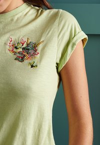 Camiseta de algodón verde claro con mangas enrolladas que presenta un bordado colorido de dragón en el pecho, destacando los detalles intrincados y la textura.