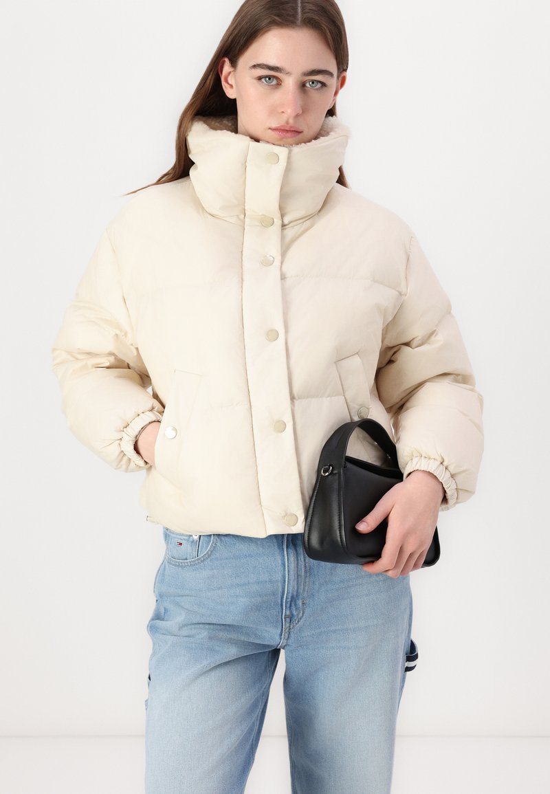 Beige Daunenjacke mit hohem Kragen und Druckknöpfen, ausgestattet mit elastischen Bündchen und zwei Vordertaschen. Kombiniert mit hellblauen Jeans und einer schwarzen Tasche.