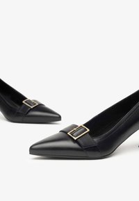 Black leather pointed-toe pump with a gold buckle accent and a medium heel. Features a smooth texture and sleek design.