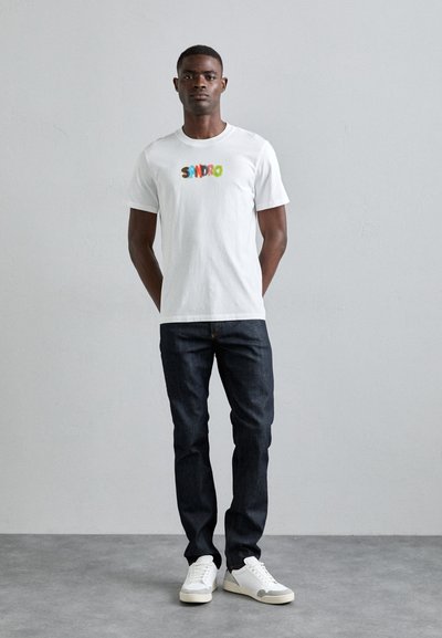 T-shirt en coton blanc avec un texte coloré "SANDRO", associé à un jean en denim noir et des baskets blanches. Look simple et décontracté.