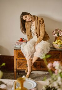 Femeie în rochie albă și jachetă maro stând pe o comodă din lemn, cu roșii, un coș cu fructe și flori într-o cameră luminată delicat.