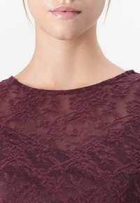 Visage et cou inférieur d'une femme portant un haut en dentelle violet translucide avec des motifs floraux sur fond blanc.