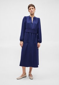 Hosszú ujjú, tengerészkék ruha függőleges rózsaszín csíkokkal, ráncolt nyakkal és övvel megkötött derékkal. Két elülső zsebe van és laza fazonú.