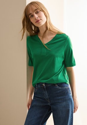 Top verde a maniche corte con scollatura a V e un morbido dettaglio di placket, abbinato a jeans blu scuro. Il tessuto appare liscio con una texture leggera.