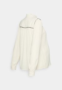 Blusa de manga longa creme com gola franzida, detalhes em preto ao longo do decote e detalhe franzido nas costas. Punhos abotoados e corte descontraído.
