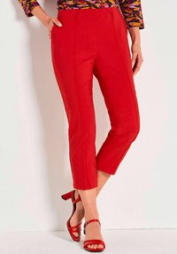 Frau in roten, kurzen Hosen und passenden roten, hohen Sandalen, steht mit einer Hand in der Tasche vor einem weißen Hintergrund.