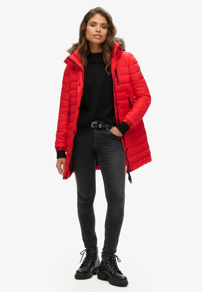 Rood gewatteerde parka met een met bont gevoerde capuchon, zwarte trui, donkere spijkerbroek en zwarte veterschoenen. Beschikt over rits- en knoopsluitingen.