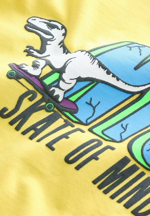 Żółty materiał z grafiką dinozaura na deskorolce, z odważnym czarnym napisem "SKATE OF MIND". Teksturowany nadruk i jasne kolory.