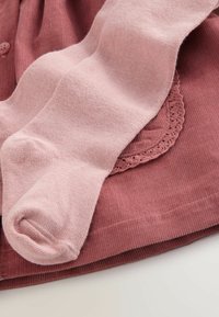 Roze katoenen panty’s zijn gerangschikt over een gestructureerde, roestkleurige ribstof. De stof heeft een gekartelde kantafwerking langs de rand.