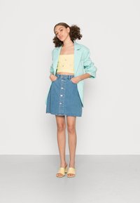 Blazer verde menta, top amarelo sem alças, saia de ganga azul com botões na frente e sapatos amarelos de salto aberto. Texturas suaves e cores vibrantes.