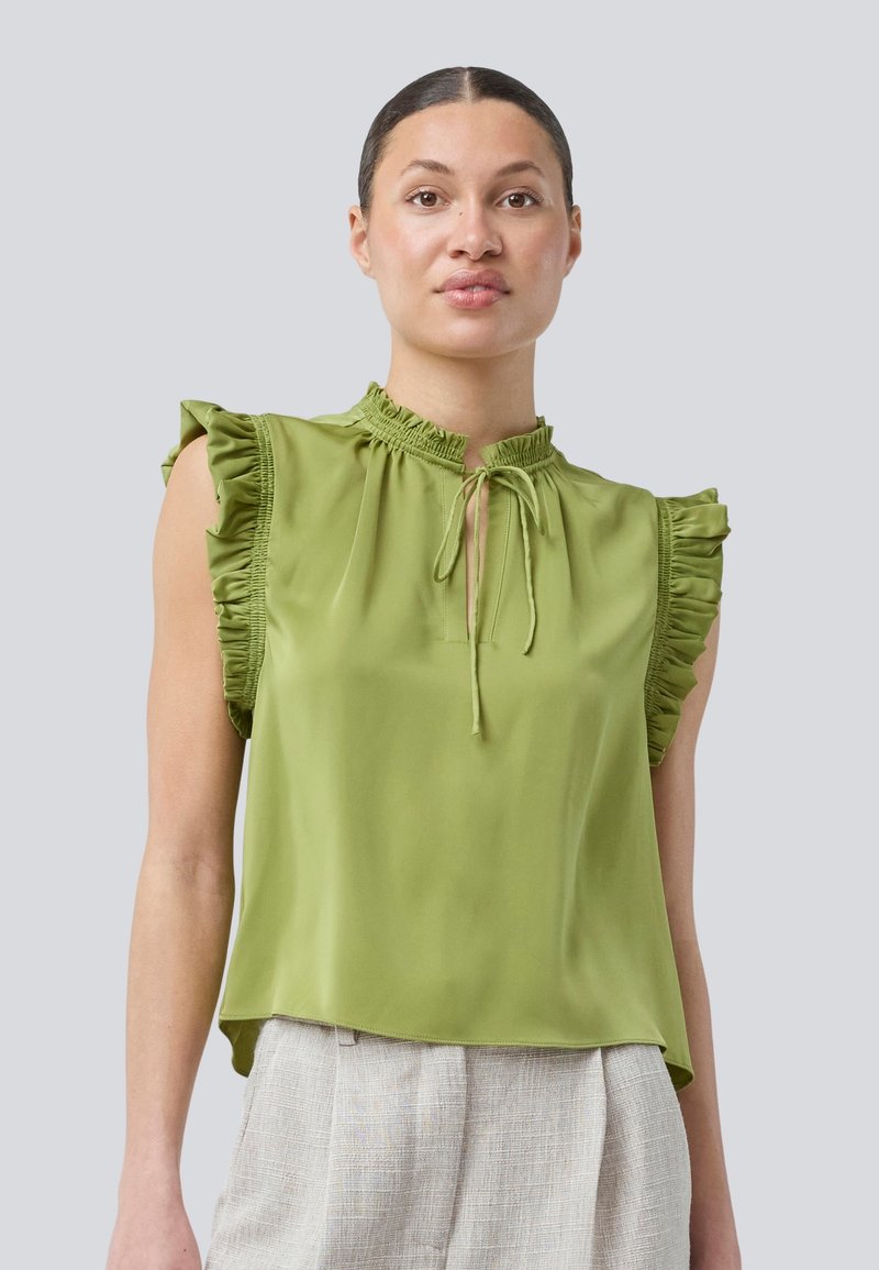 Green blouse with frilled sleeves, high neck, and a tie detail. Smooth fabric with a relaxed fit, worn with light-colored textured pants.