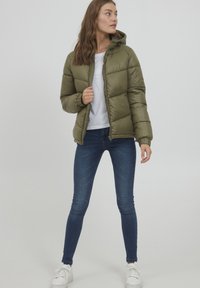 Giacca puffer verde oliva con cappuccio, chiusura con zip, texture trapuntata, abbinata a jeans attillati blu scuri e sneaker bianche.
