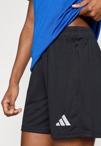 Μαύρο αθλητικό σορτς με ελαστική μέση, πλαϊνή τσέπη και λευκό λογότυπο Adidas στο κάτω αριστερό μέρος. Το ύφασμα φαίνεται λείο και ελαφρύ.