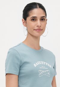 Šviesiai mėlyna trumpomis rankovėmis marškinėliai, su baltu atspaudu, kuriame pavaizduoti kryžiumi sudėti bagetės ir tekstas "BAGUETTES DE LA BOULANGERIE."