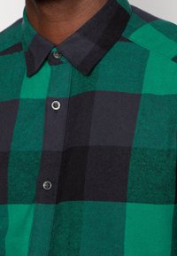 Primer plano de una camisa de franela a cuadros verdes y negros abotonada en el cuello, usada por una persona de piel oscura.