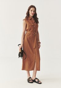Vestido sin mangas marrón con botones frontales, cintura con cinturón y dos bolsillos en el pecho. Combinado con sandalias negras y un bolso pequeño.