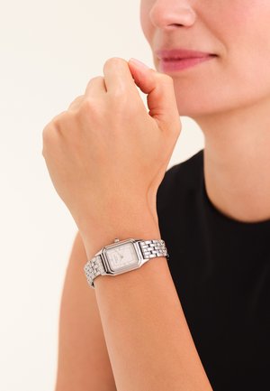 Zilveren metalen polshorloge met een rechthoekige witte wijzerplaat en gestructureerde band, gedragen om een licht huidtint pols tegen een effen achtergrond.