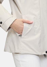 Veste imperméable beige clair avec une poche zippée et des détails de poignet boutonnés. Présente une texture lisse et une coupe ajustée.