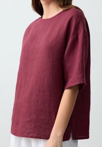 Femme portant un haut ample en lin bordeaux à manches courtes avec des fentes latérales, visible du cou jusqu'aux hanches sur un fond uni.