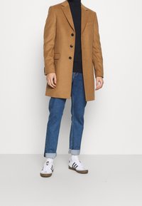 Maschio che indossa un cappotto marrone con bottoni, dolcevita nero, jeans blu risvoltati, calzini bianchi e sneaker bianche con strisce nere.