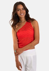 Top rojo asimétrico hecho de tela texturizada, con rayas horizontales. Se lleva con pantalones blancos, complementado con pulseras en capas.