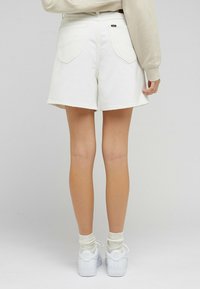 Bílé džíny šortky s třepeným lemem, dvěma zadními kapsami a vypasovaným pasem. Nosič má bílé tenisky a strukturované bílé ponožky.